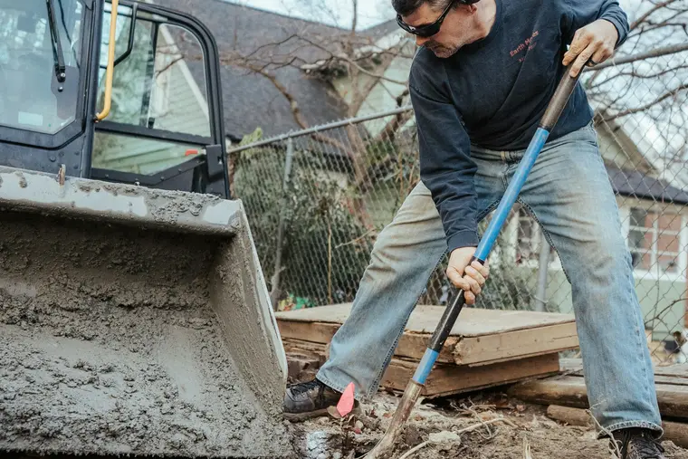 Worker with a shovel beside a mini-excavator bucket in a residential yard